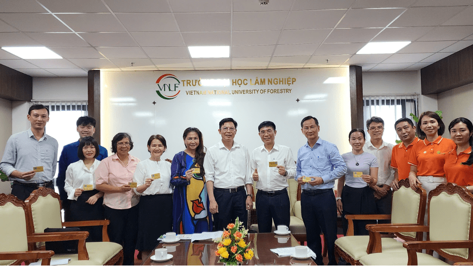 Một nhóm người đứng chụp ảnh bên trong Trường Đại học Lâm nghiệp, với logo của trường trên tường. Mọi người đều mỉm cười và cầm thẻ trong tay.