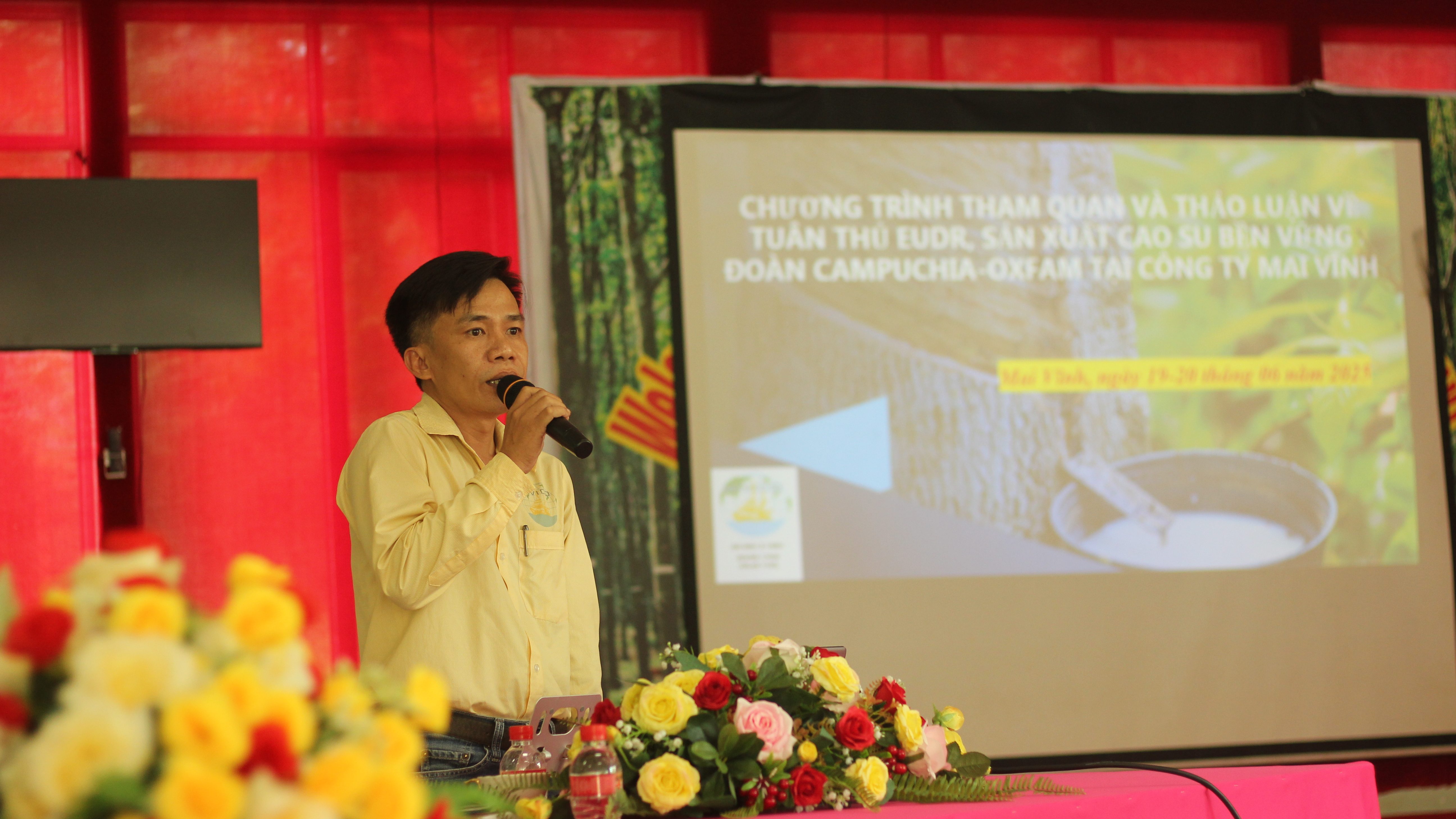 Một người đàn ông đang nói chuyện trước khán giả trong một sự kiện, với phông nền là bảng thông tin về chương trình tham quan và thảo luận liên quan đến EUDR tại Công ty Mai Vĩnh.