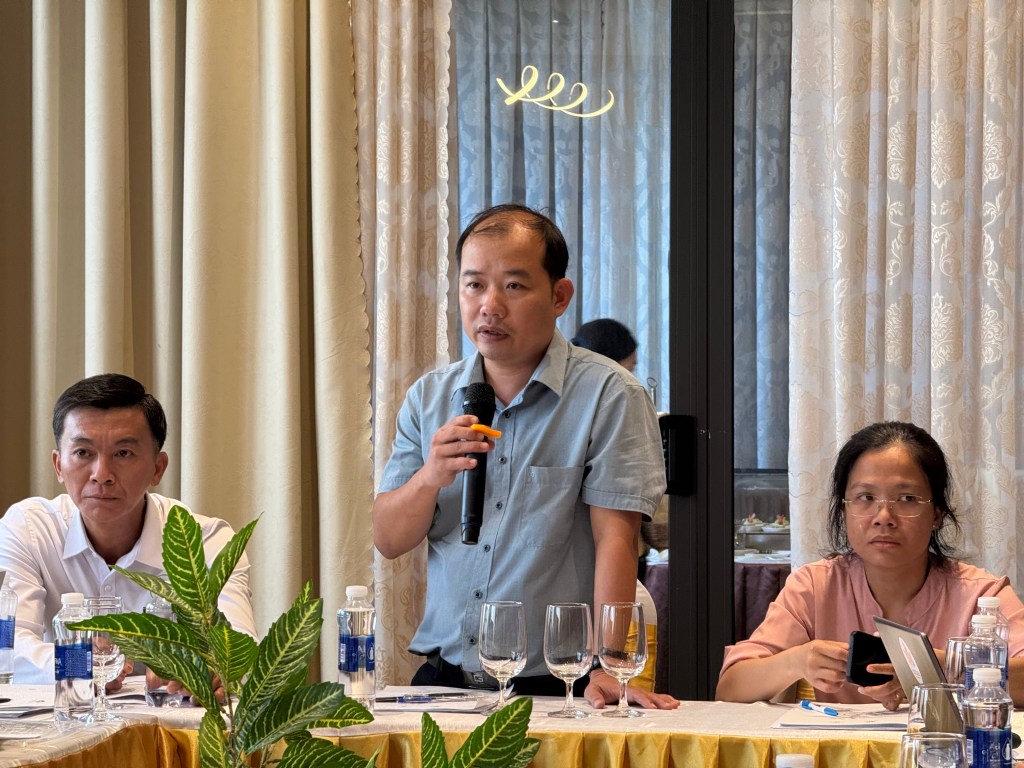 Người đàn ông đang nói chuyện tại cuộc họp với các thành viên khác ngồi lắng nghe, trong bối cảnh một hội trường có rèm cửa và bàn ghế.