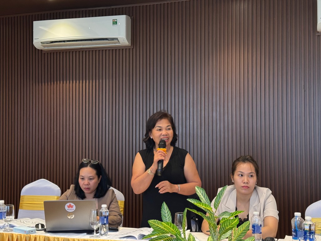 Một người phụ nữ đang phát biểu tại cuộc họp bàn về Sổ tay hướng dẫn cho hộ cao su tiểu điền và đại lý, phía sau là các người tham dự và bàn họp có máy tính và nước uống.