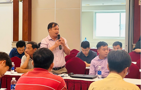 Một người đàn ông đang nói trong một buổi hội thảo, xung quanh là nhiều người tham gia lắng nghe.