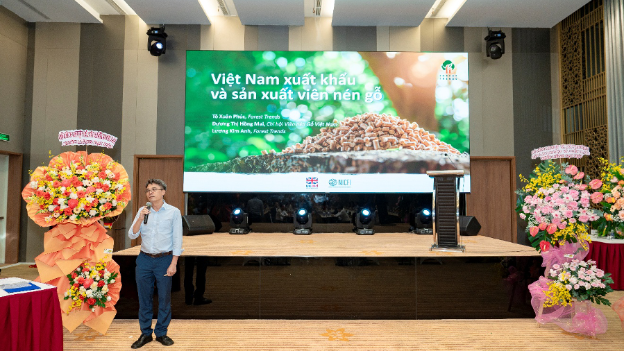 Một đại biểu đang phát biểu tại sự kiện về xuất khẩu và sản xuất viên nén gỗ Việt Nam, với màn hình lớn phía sau trình bày thông tin liên quan. Bên cạnh có hoa chúc mừng được sắp xếp trang trí.